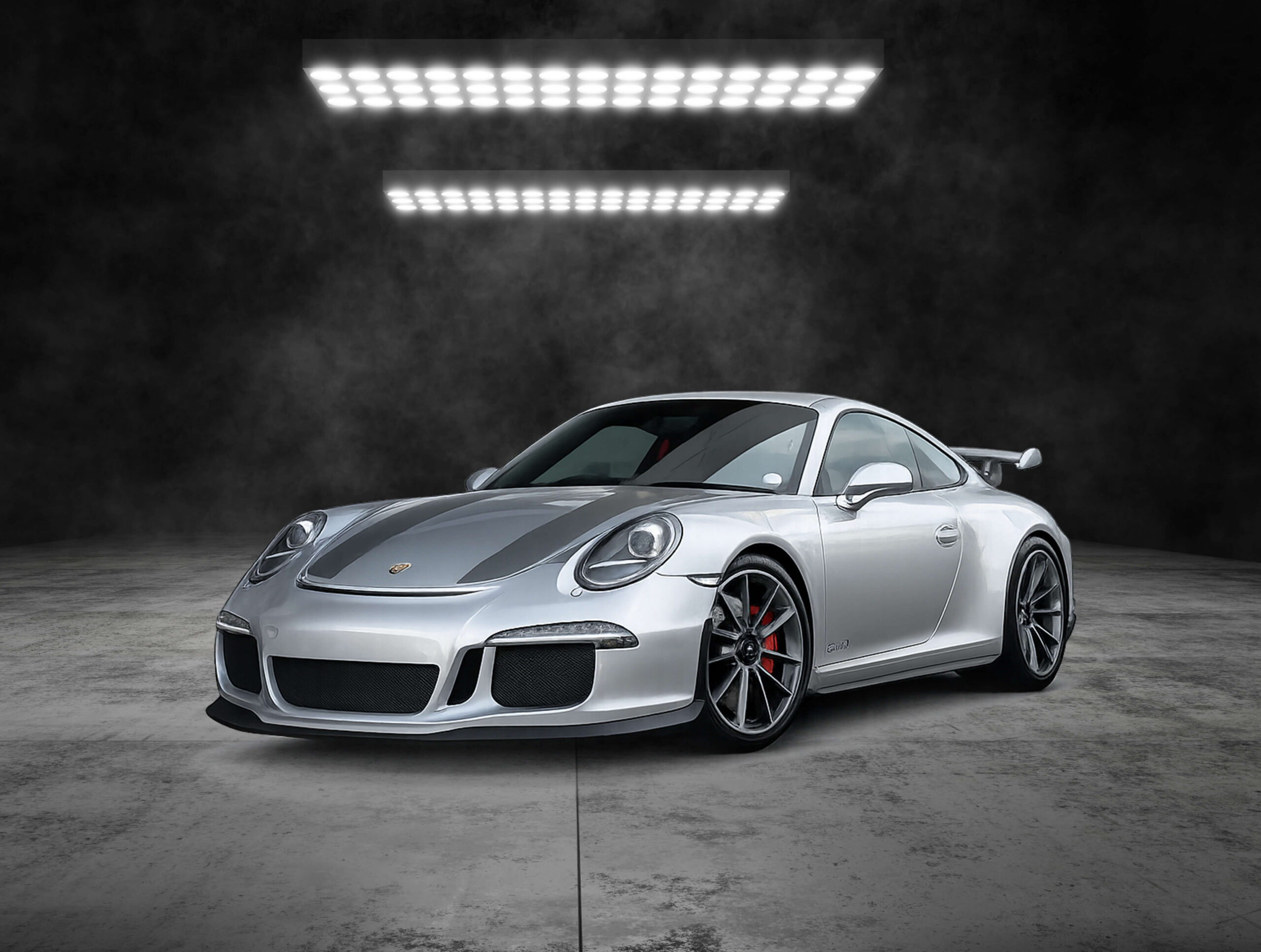 Front three-quarter view of a silver Porsche sports car with a dark center stripe, set against a dark smoky studio backdrop and bright overhead lights.